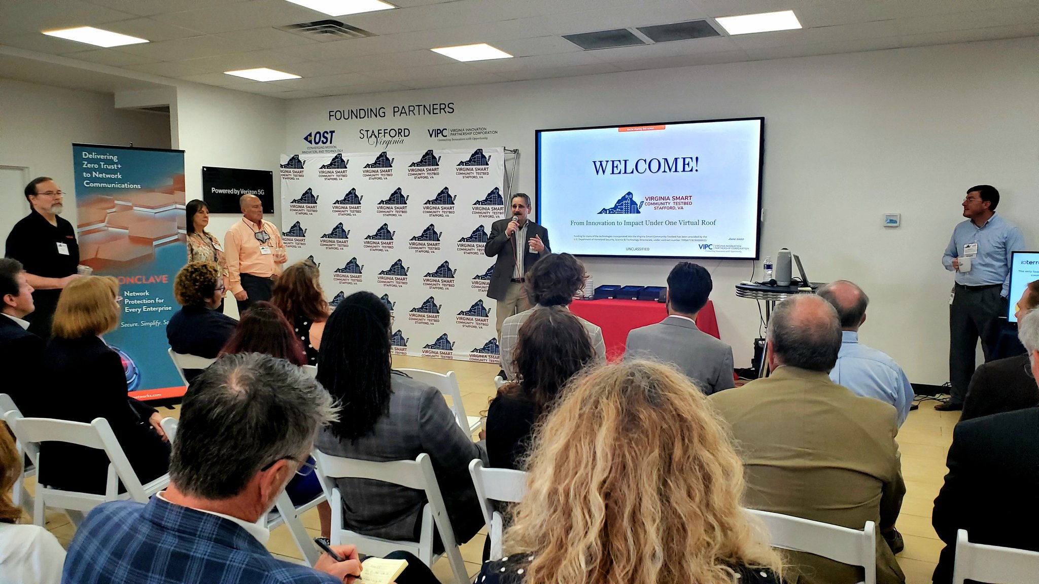 Room with attendees, most sitting with few standing dress in business casual attire, are watching a man talking in to a microphone, presenting information shown on a big screen tv.